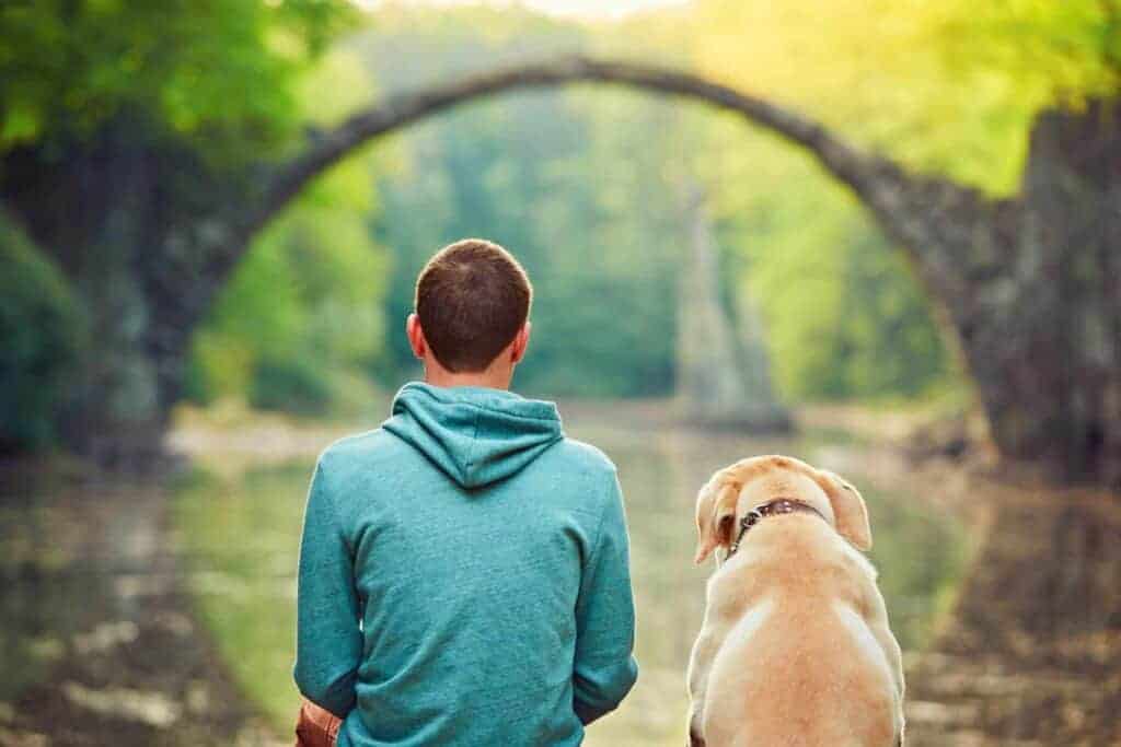 Nachdenklicher junger Mann sitzt mit seinem Hund am Ufer eines Teiches